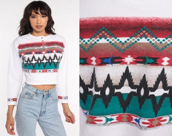 May include: White long-sleeve shirt with a cropped length and a geometric pattern in red, brown, green, and black. The pattern is reminiscent of Native American designs.