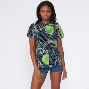 May include: A short-sleeved top with a navy blue background and a pattern of green, yellow, and white swirls and lines. The top is paired with denim shorts. The model is standing in front of a white background.