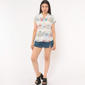 May include: A short-sleeved white button-down shirt featuring a colorful hand-drawn style print of city buildings and street scenes, paired with a dark blue denim mini skirt that has a small side slit and black platform sandals.