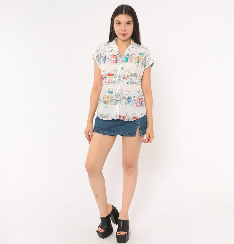 May include: A short-sleeved white button-down shirt featuring a colorful hand-drawn style print of city buildings and street scenes, paired with a dark blue denim mini skirt that has a small side slit and black platform sandals.