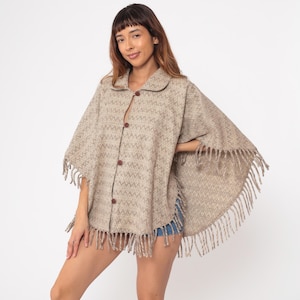 May include: A beige poncho with a geometric pattern and fringe trim. The poncho has a collared neckline and is fastened with brown buttons down the front. The fringe hangs along the bottom edge and sleeves. The garment is made of a woven fabric.