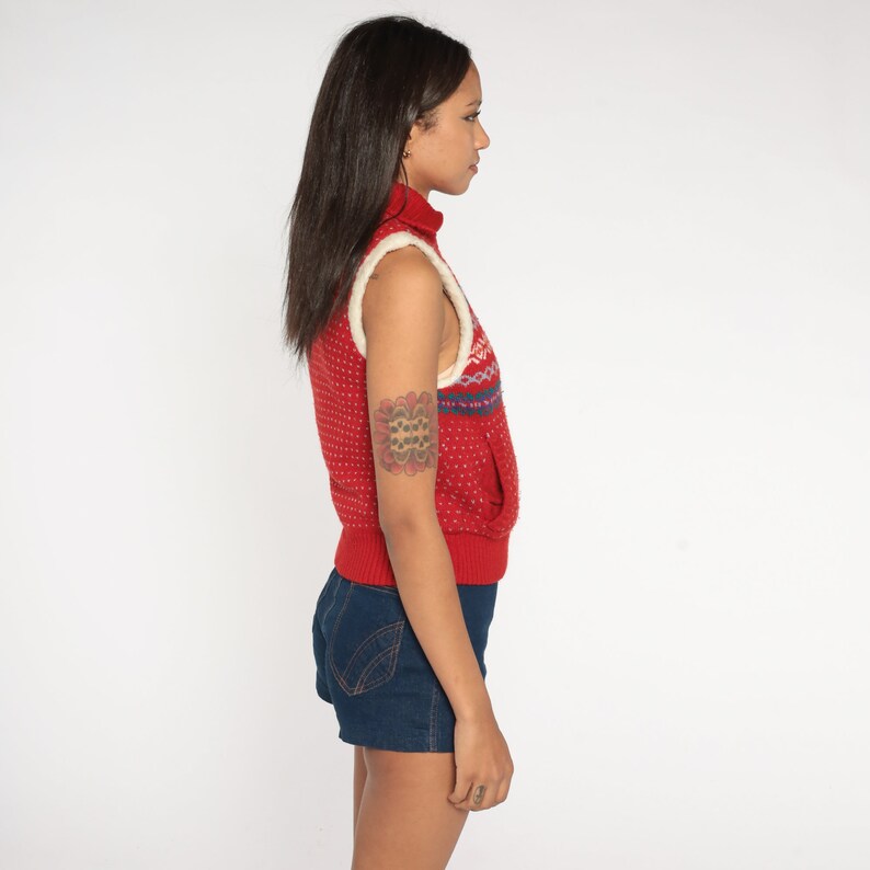 May include: Red and white patterned sleeveless sweater vest with a mock neck and a white trim. The vest is made of a soft, knitted fabric.