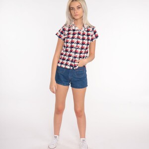 May include: A white short-sleeved button-up shirt with a red and blue geometric pattern. The shirt has a collar and a pocket on the left side. The model is wearing blue denim shorts and white sneakers.