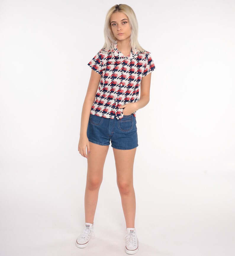 May include: A white short-sleeved button-up shirt with a red and blue geometric pattern. The shirt has a collar and a pocket on the left side. The model is wearing blue denim shorts and white sneakers.
