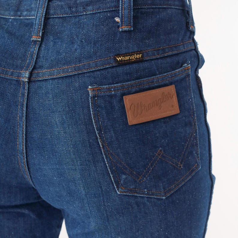 May include: Close-up of dark blue denim jeans. The back pocket features a brown leather patch with the brand name "Wrangler" embossed. The jeans have classic stitching details and a small "Wrangler" label above the back pocket.
