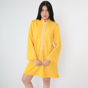 May include: A yellow long-sleeved dress with a white trim down the front. The dress has a high collar and a loose fit.