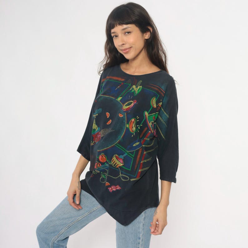 May include: A dark navy blue tunic-style top with a colorful abstract design. The three-quarter sleeve shirt features a round neckline and a unique asymmetrical hemline. The artwork includes red, green, and yellow shapes on a dark background.