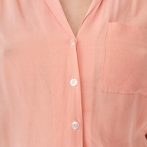 Puede incluir: Primer plano de una camisa abotonada de color melocot&oacute;n. La camisa tiene un cuello cl&aacute;sico, un bolsillo en el pecho y tres botones blancos visibles. La tela parece suave y lisa.