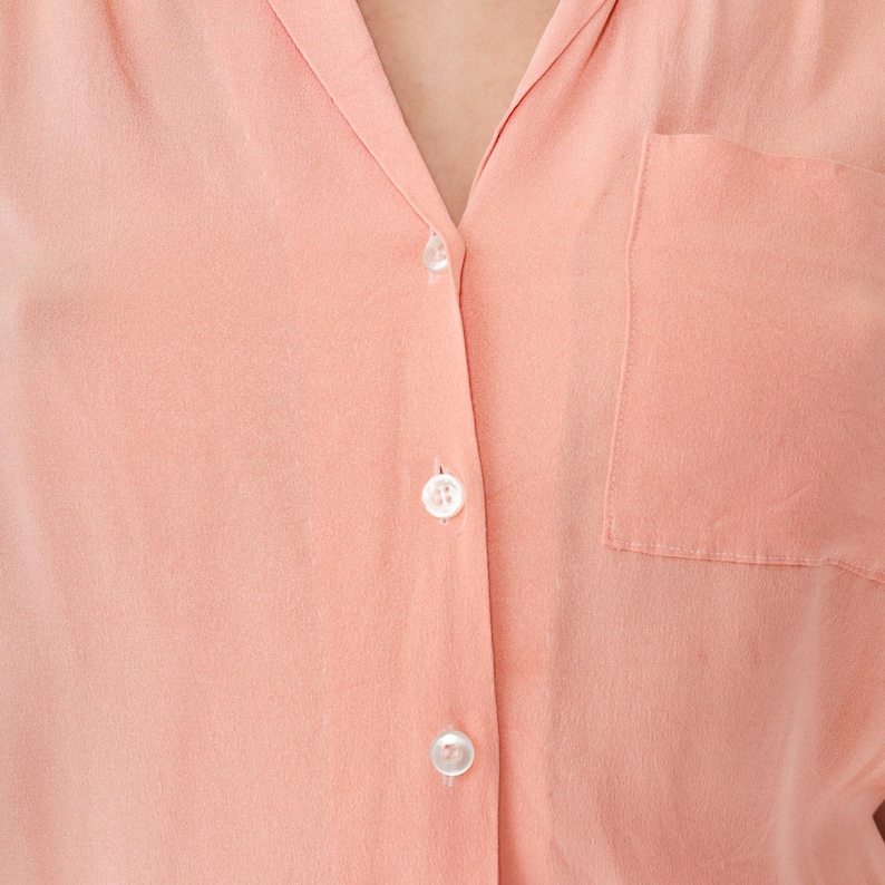 Puede incluir: Primer plano de una camisa abotonada de color melocot&oacute;n. La camisa tiene un cuello cl&aacute;sico, un bolsillo en el pecho y tres botones blancos visibles. La tela parece suave y lisa.