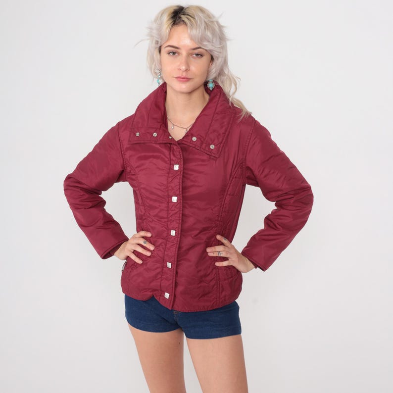 May include: A burgundy quilted jacket with a high collar and silver snap closures. The jacket has long sleeves and a fitted silhouette. The model is wearing blue denim shorts. The jacket is a vintage style.