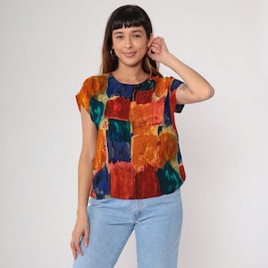 May include: A short-sleeved top with a colorful abstract pattern. The top features a patchwork design with squares of red, orange, blue, and green. The top is worn with light blue jeans.