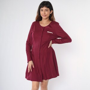 May include: A burgundy button-down dress with long sleeves and a slightly flared skirt. The dress has a small white embellishment on the left side of the chest.