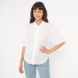 May include: A white button-down shirt with short sleeves. The shirt has a classic collar and is paired with light blue jeans. The model is smiling and has dark hair.