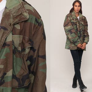 May include: A camouflage field jacket featuring a woodland pattern in shades of olive green, brown, and black. The jacket includes a high collar, a front zipper closure with metal snap buttons, and two chest pockets with flaps over the pockets.