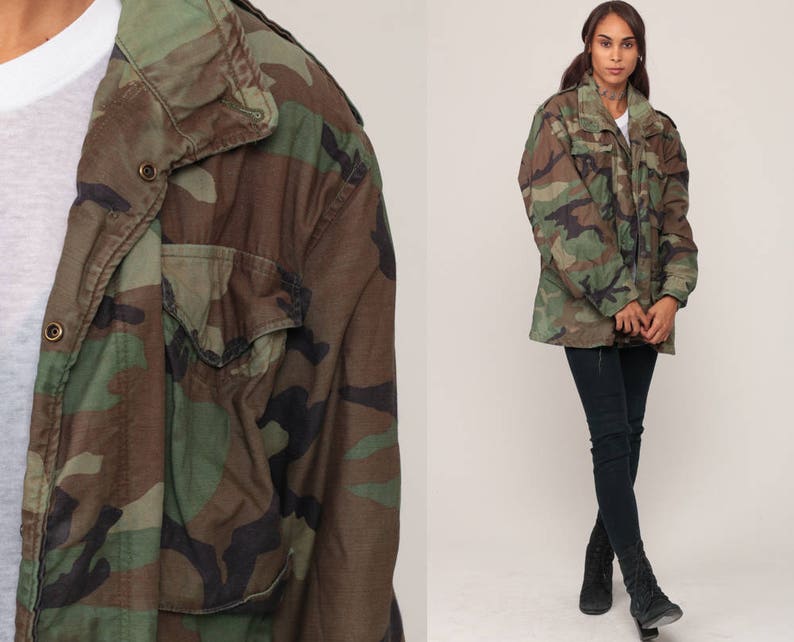 May include: A camouflage field jacket featuring a woodland pattern in shades of olive green, brown, and black. The jacket includes a high collar, a front zipper closure with metal snap buttons, and two chest pockets with flaps over the pockets.