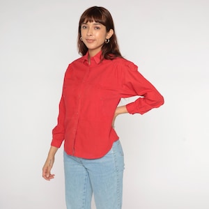 May include: A bright red button-down shirt with long sleeves and two chest pockets. The shirt is paired with light blue denim jeans. The model is standing in front of a white background.