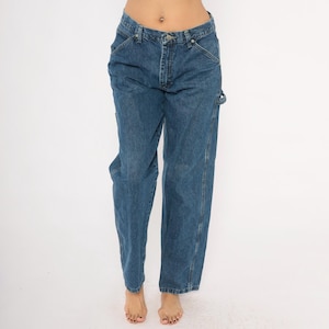 May include: Blue denim jeans with a classic five-pocket design. The jeans have a straight leg fit and feature a button and zipper closure. The jeans have a tool pocket on the right leg. The jeans are worn by a person.