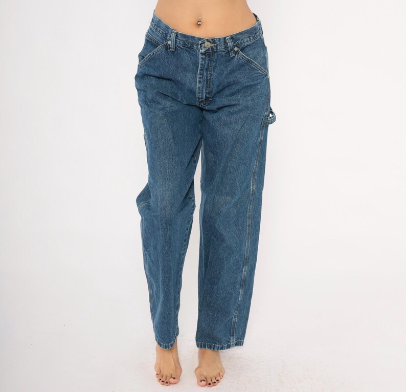 May include: Blue denim jeans with a classic five-pocket design. The jeans have a straight leg fit and feature a button and zipper closure. The jeans have a tool pocket on the right leg. The jeans are worn by a person.