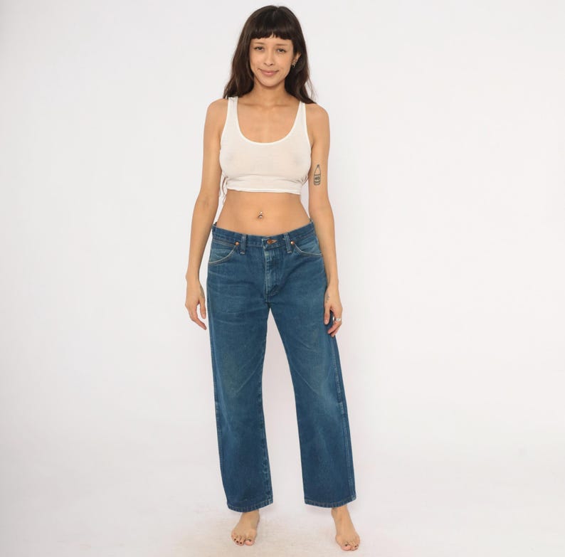 May include: A woman wearing a white tank top and blue jeans. The jeans are a classic, straight-leg style in a medium wash denim. The tank top is a light, neutral color, and the overall look is casual and simple.