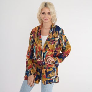 May include: A colorful blazer with an abstract pattern in shades of blue, red, yellow, and green. The blazer has a relaxed fit, long sleeves, and two front pockets. The garment is unbuttoned, revealing a white top underneath.