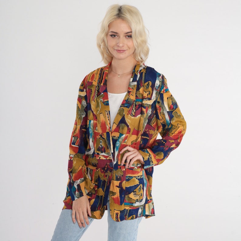 May include: A colorful blazer with an abstract pattern in shades of blue, red, yellow, and green. The blazer has a relaxed fit, long sleeves, and two front pockets. The garment is unbuttoned, revealing a white top underneath.