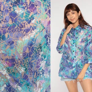 Op de afbeelding: Een overhemd met knopen en een levendig, abstract patroon in tinten blauw, paars en teal. De stof heeft een semi-transparante kwaliteit. Het overhemd heeft een klassieke kraag en driekwartmouwen.
