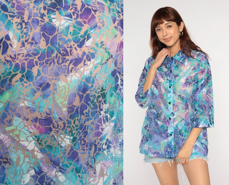 Op de afbeelding: Een overhemd met knopen en een levendig, abstract patroon in tinten blauw, paars en teal. De stof heeft een semi-transparante kwaliteit. Het overhemd heeft een klassieke kraag en driekwartmouwen.