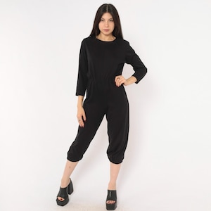 May include: A solid black jumpsuit featuring a round neckline, three-quarter length sleeves, and a cinched waistline. The garment has a relaxed fit through the legs, which taper down to elasticized cuffs at the mid-calf, paired with black platform sandals.
