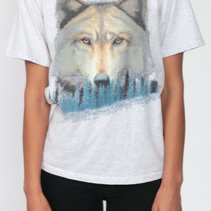 May include: A gray t-shirt with a wolf graphic. The wolf is facing forward with its eyes open and its mouth closed. The wolf is surrounded by a blue and green forest.