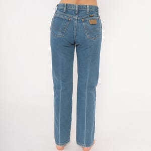 May include: Blue denim jeans with a classic five-pocket design. The jeans feature a straight leg cut and a brown leather patch on the back pocket. The Wrangler brand is visible on the patch.