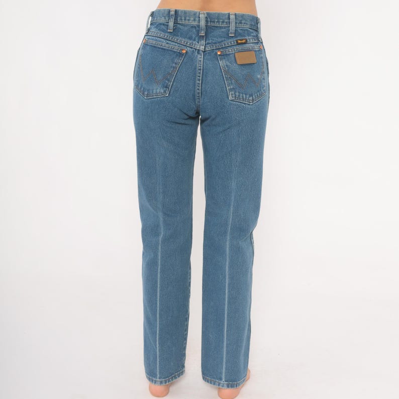 May include: Blue denim jeans with a classic five-pocket design. The jeans feature a straight leg cut and a brown leather patch on the back pocket. The Wrangler brand is visible on the patch.
