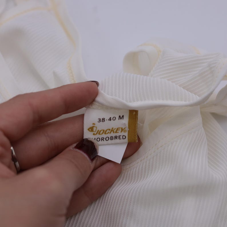 Peut inclure: Gros plan sur un v&ecirc;tement blanc c&ocirc;tel&eacute; avec une &eacute;tiquette. L'&eacute;tiquette indique "38-40 M" et pr&eacute;sente le logo "Jockey" et le mot "Thoroughbred". Le v&ecirc;tement est fait d'un tissu doux et de couleur claire.
