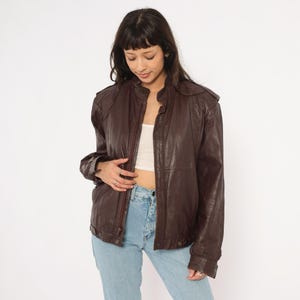 May include: A brown leather jacket with a zip-up front, shoulder pads, and a stand-up collar. The jacket has a slightly worn appearance and is paired with light blue jeans. The jacket is a vintage style.