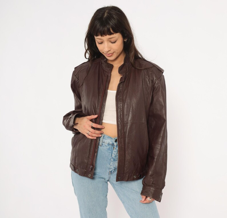 May include: A brown leather jacket with a zip-up front, shoulder pads, and a stand-up collar. The jacket has a slightly worn appearance and is paired with light blue jeans. The jacket is a vintage style.