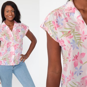 May include: A short-sleeved button-down shirt with a floral pattern. The shirt features a white background with pink, blue, and green floral designs. The shirt has a collar and is made of a lightweight fabric.