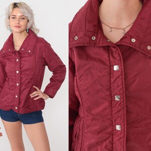 May include: A burgundy quilted jacket with a high collar and silver square buttons. The jacket has a fitted waist and long sleeves. The model is wearing denim shorts. The jacket is a vintage style.