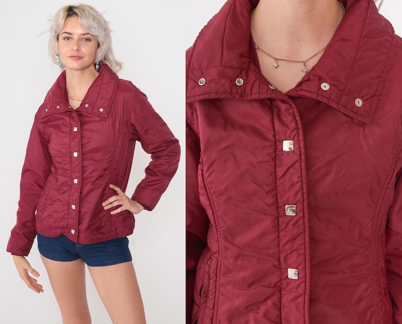 May include: A burgundy quilted jacket with a high collar and silver square buttons. The jacket has a fitted waist and long sleeves. The model is wearing denim shorts. The jacket is a vintage style.