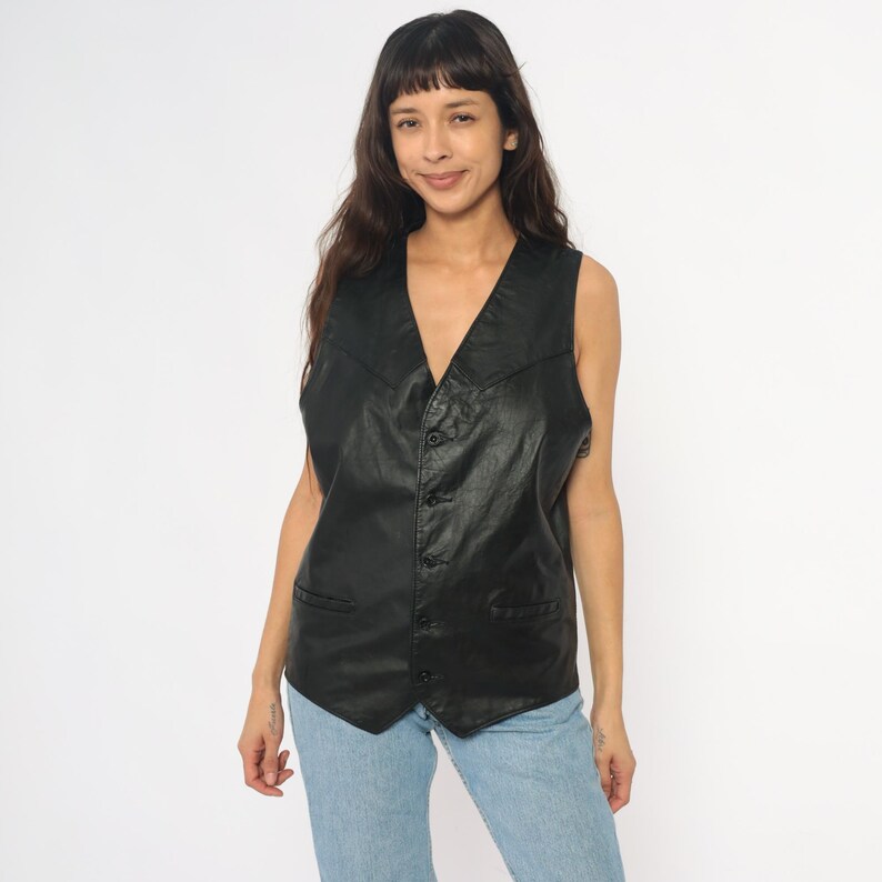 May include: A black leather vest with a V-neck and button closure. The vest has two front pockets and a tailored fit. The model is wearing the vest with blue jeans. The vest is a classic style.