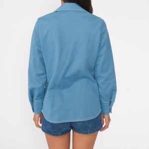 May include: A light blue long-sleeved shirt with a white stitch detail around the collar and cuffs. The shirt is buttoned up and has a relaxed fit.