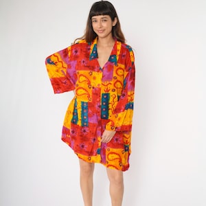 Könnte beinhalten: Ein farbenfrohes, gemustertes Hemdkleid mit Knopfleiste und langen Ärmeln. Das Kleid hat ein Patchwork-Design in Rot, Gelb, Blau und Lila.
