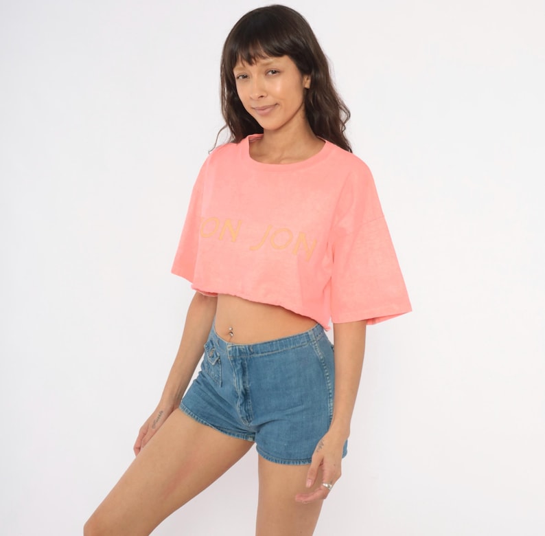 K&ouml;nnte beinhalten: Ein korallenfarbenes, bauchfreies T-Shirt mit kurzen &Auml;rmeln und Rundhalsausschnitt, das auf der Brust den Schriftzug "BON JON" in einem helleren Rosaton aufweist, kombiniert mit blauen Jeansshorts mit Knopfleiste und einer kleinen Vordertasche.