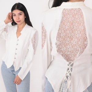 May include: A white blouse with long sleeves, a V-neck, and decorative buttons. The blouse has lace detailing on the sleeves and back, with a corset-style lace-up closure. The design blends classic and romantic elements.