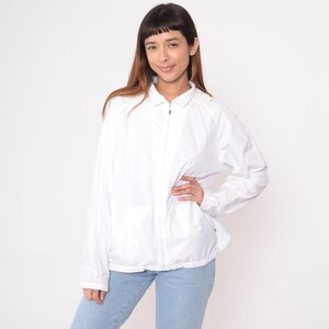 May include: A white, lightweight jacket with a full zipper closure. The jacket has long sleeves and a collared neckline. The person is wearing light blue jeans. The background is plain white.