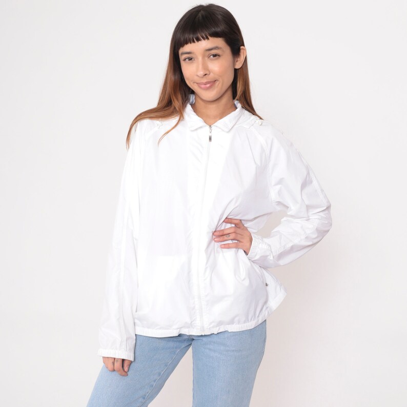 May include: A white, lightweight jacket with a full zipper closure. The jacket has long sleeves and a collared neckline. The person is wearing light blue jeans. The background is plain white.