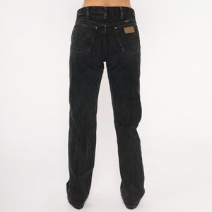 May include: Black denim Wrangler jeans. The jeans have a classic five-pocket design, a straight leg, and a brown leather patch on the back. The jeans are worn and have a faded appearance.