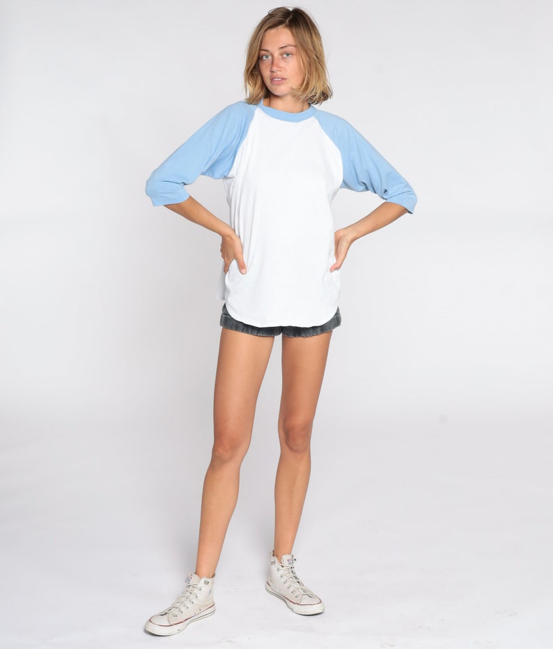 May include: A white baseball tee with light blue sleeves. The shirt is paired with black denim shorts and high-top canvas sneakers. The model is standing with their hands on their hips against a white backdrop.