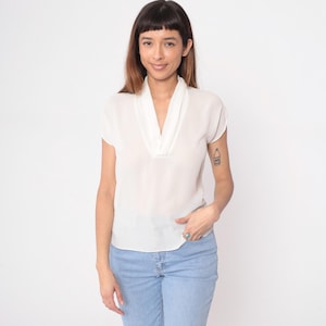 Puede incluir: Una blusa color crema con cuello en V y mangas cortas. La blusa se combina con vaqueros azul claro. La mujer de la imagen sonríe y tiene un tatuaje en el brazo.