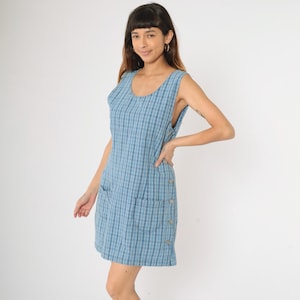 Könnte beinhalten: Ein ärmelloses Kleid in blau-weißem Karomuster mit Rundhalsausschnitt und zwei Taschen. Das Kleid hat einen seitlichen Knopfverschluss.