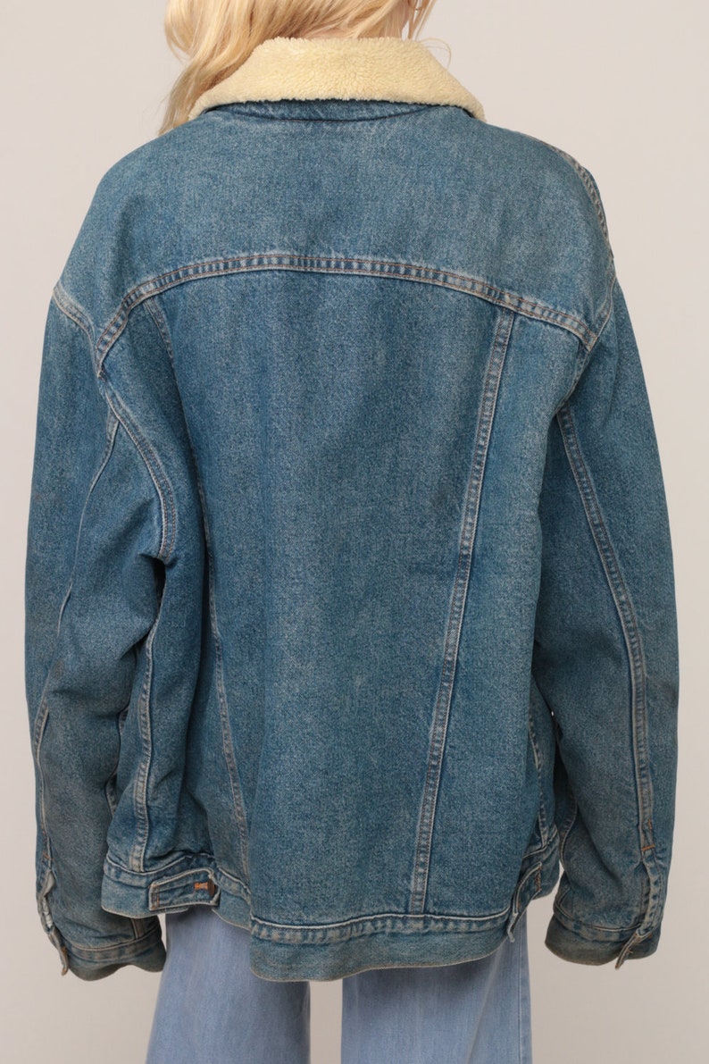 Puede incluir: Una chaqueta de mezclilla azul con un cuello de sherpa de color crema. La chaqueta tiene mangas largas, un canes&uacute; en la espalda y detalles de costuras verticales. La tela de mezclilla tiene un lavado azul medio con costuras visibles.
