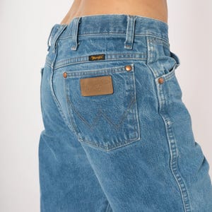 Puede incluir: Vaqueros de mezclilla azul claro con un dise&ntilde;o cl&aacute;sico de cinco bolsillos. Cuenta con un parche de cuero marr&oacute;n con el logotipo "Wrangler" en el bolsillo trasero. Los vaqueros tienen remaches de cobre y corte recto.